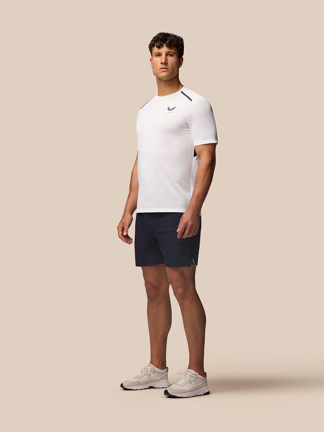 Male model wearing white Apex Training Short Sleeve T Shirt with navy shorts against a beige background.
