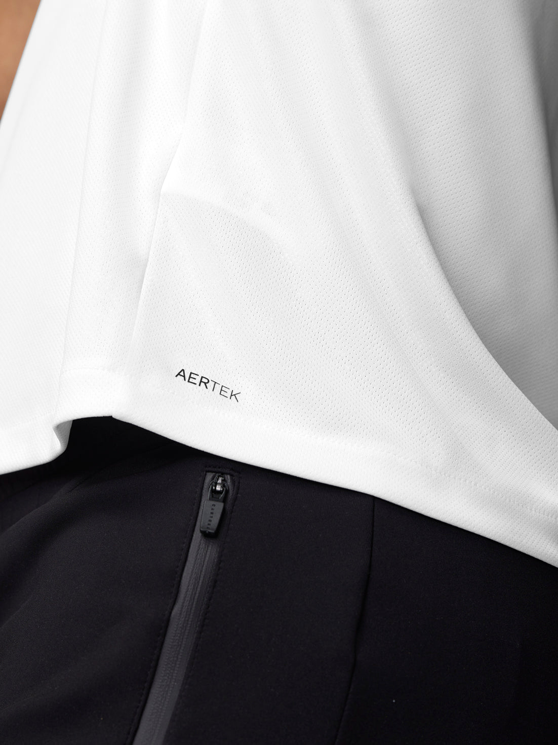 Close up of white training T-shirt hem showing Aertek branding with black pants zipper nearby
