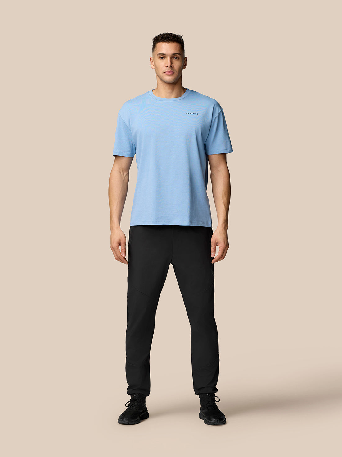 Full-body shot of a man wearing black Flex joggers and a light blue t-shirt against a beige background
