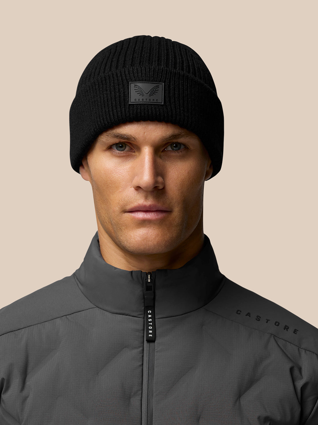 Head and shoulders shot of model in black ribbed Castore beanie with front patch and grey jacket