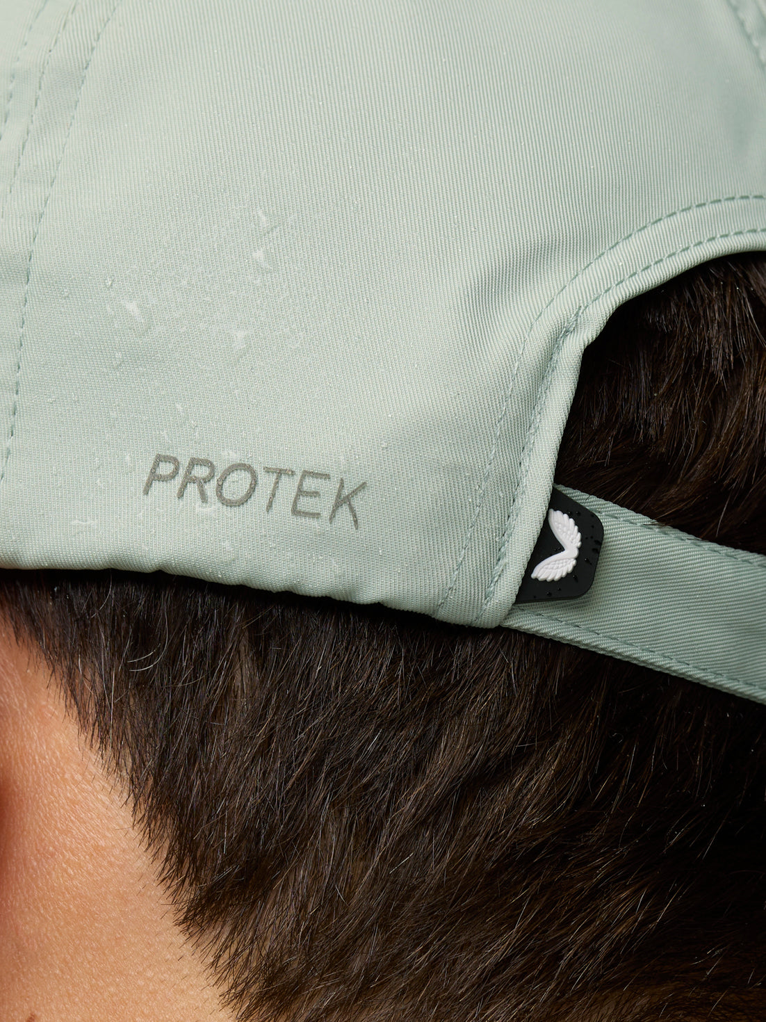 Close-up of the back of a sage cap showing PROTEK branding on the strap and a small winged badge