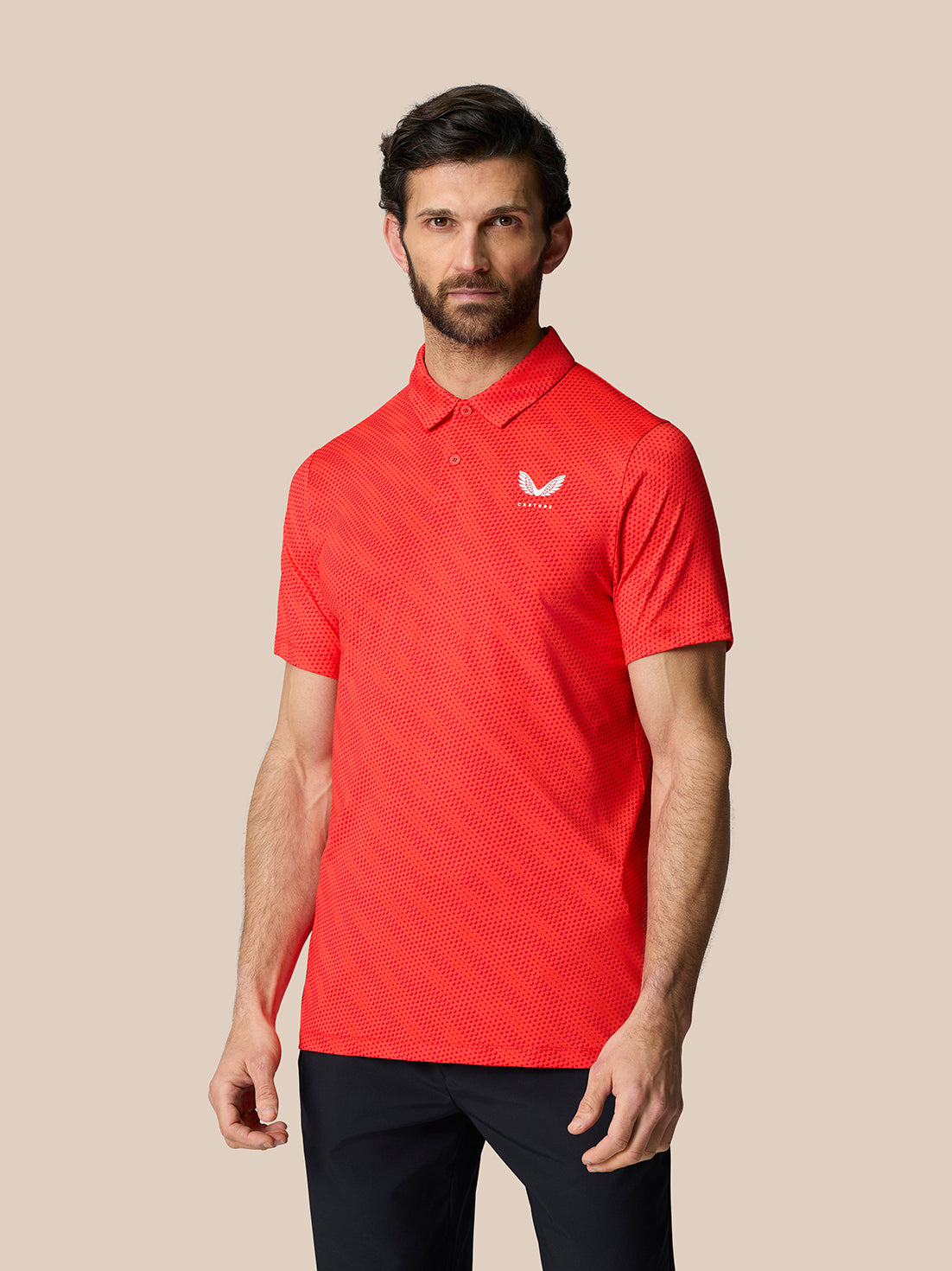 Male model in red Club Graphic Polo with golf ball pattern and white Castore logo on chest standing against a beige background