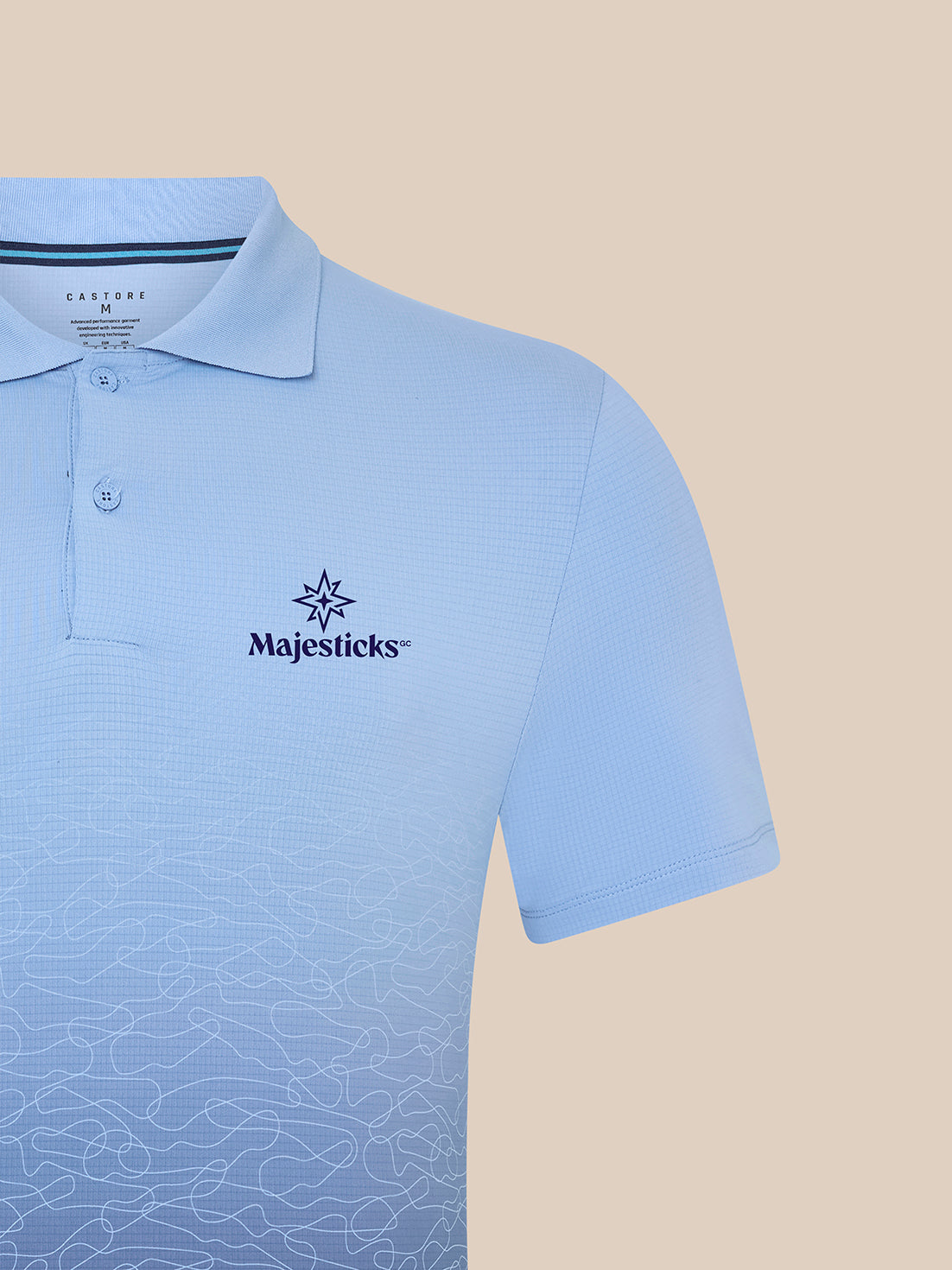 Close-up of sky blue Majesticks golf polo showing left chest logo and subtle ombre print across the lower front.
