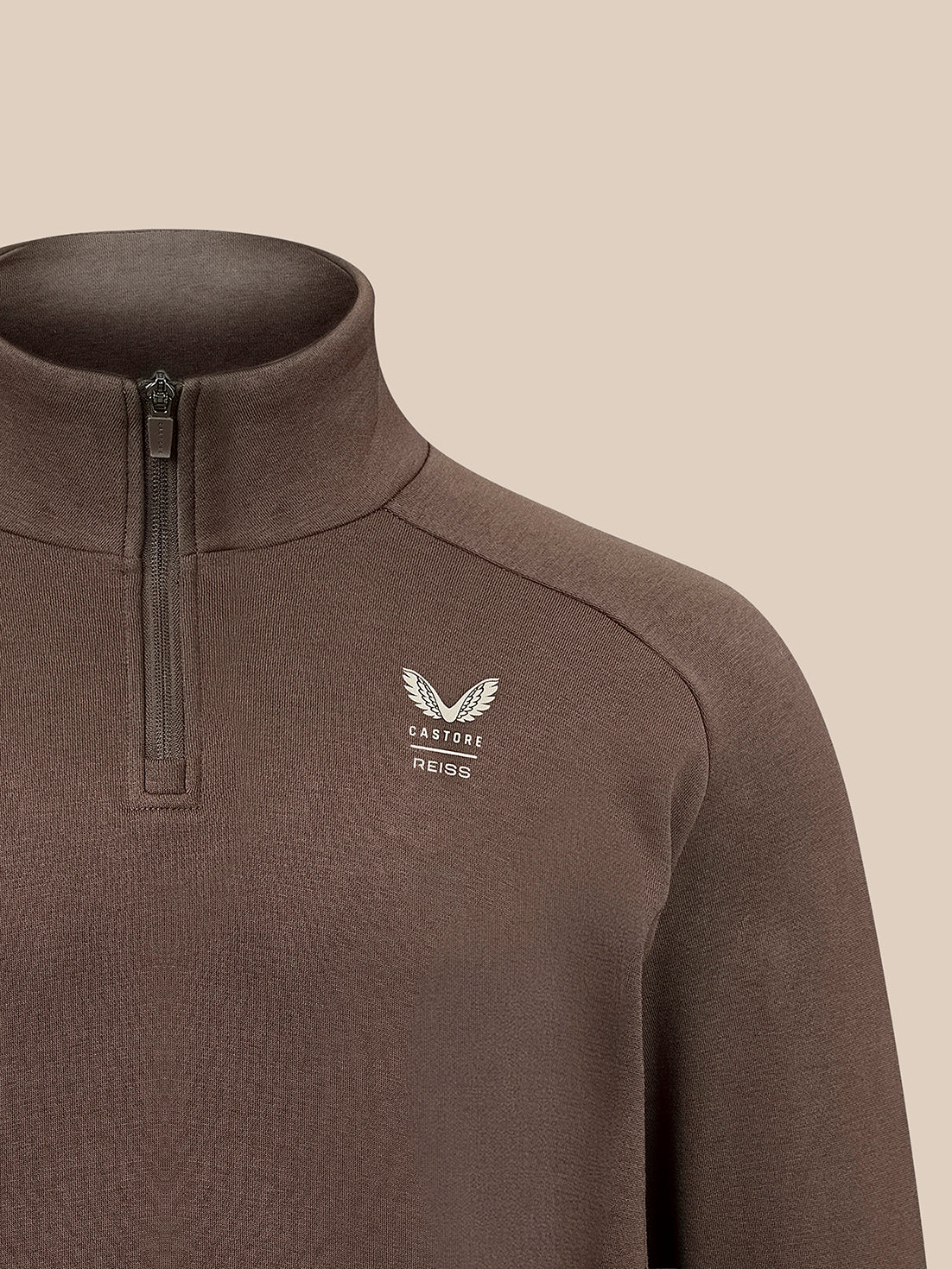 A close-up of a dark brown quarter-zip pullover sweater with a white embroidered winged logo and the brand name 'CASTORE' on the left chest, set against a solid beige background.