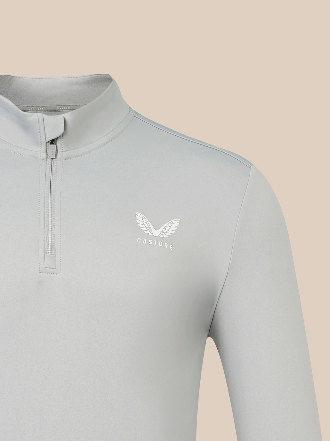 A close-up of a light gray long-sleeve quarter-zip pullover with a white winged logo and the word 'CASTORE' on the left chest, set against a solid beige background.