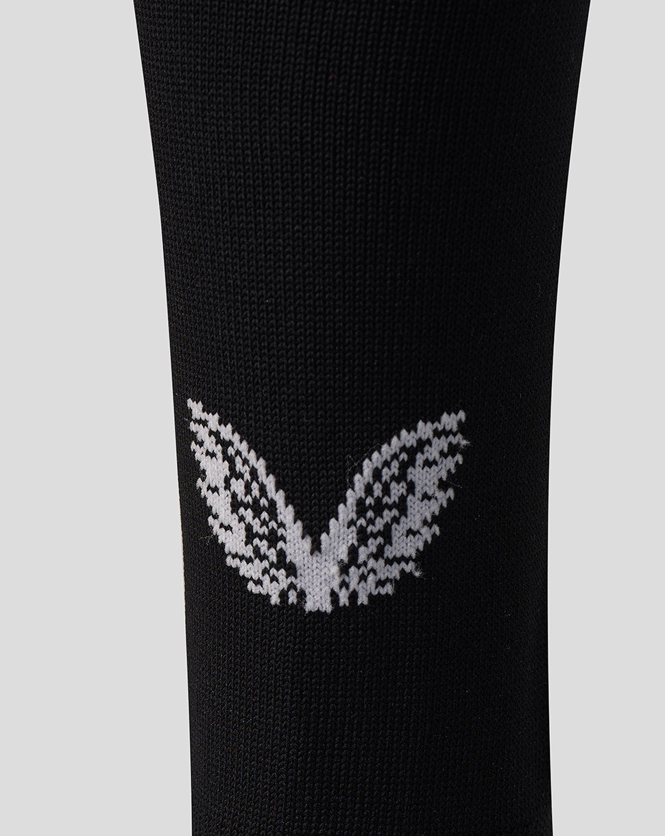 A close-up of a black knit sock featuring a white knitted butterfly motif on the lower calf, set against a solid light gray background.