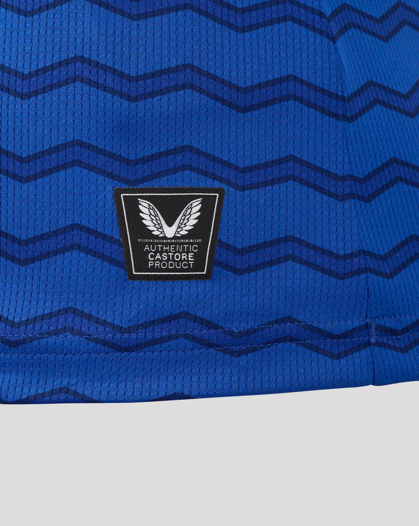 Close up of blue Everton women's 25/26 home shirt fabric with dark chevron waves and a black patch featuring white wings and Authentic Castore Product text.