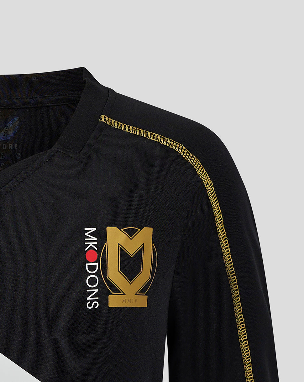 Close-up of chest on black MK DONS junior 24/25 third shirt showing a large gold crest and vertical MK DONS lettering beside it with gold shoulder seam.