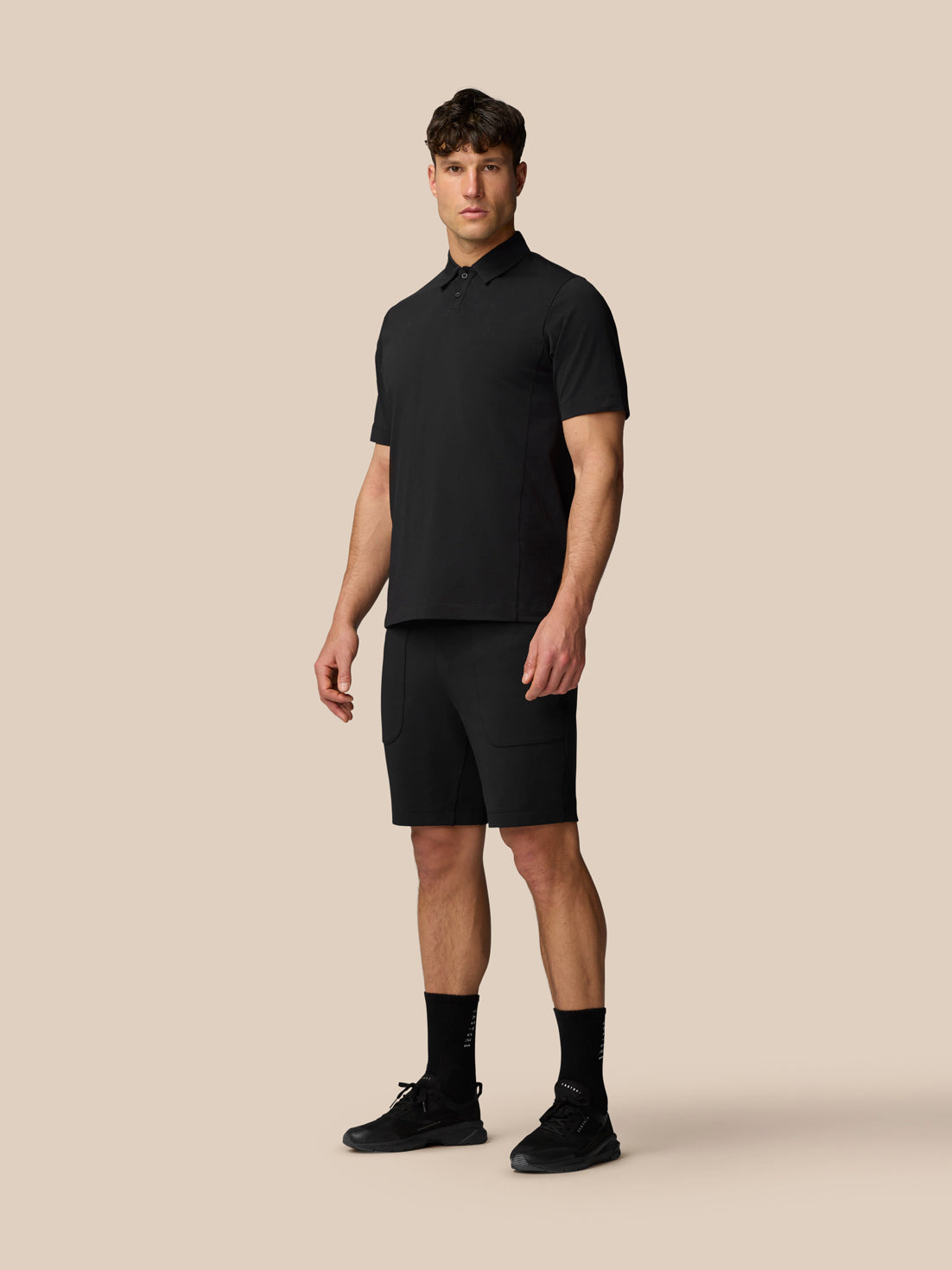 Full-body view of a man in black travel shorts and a black polo shirt against a beige background.