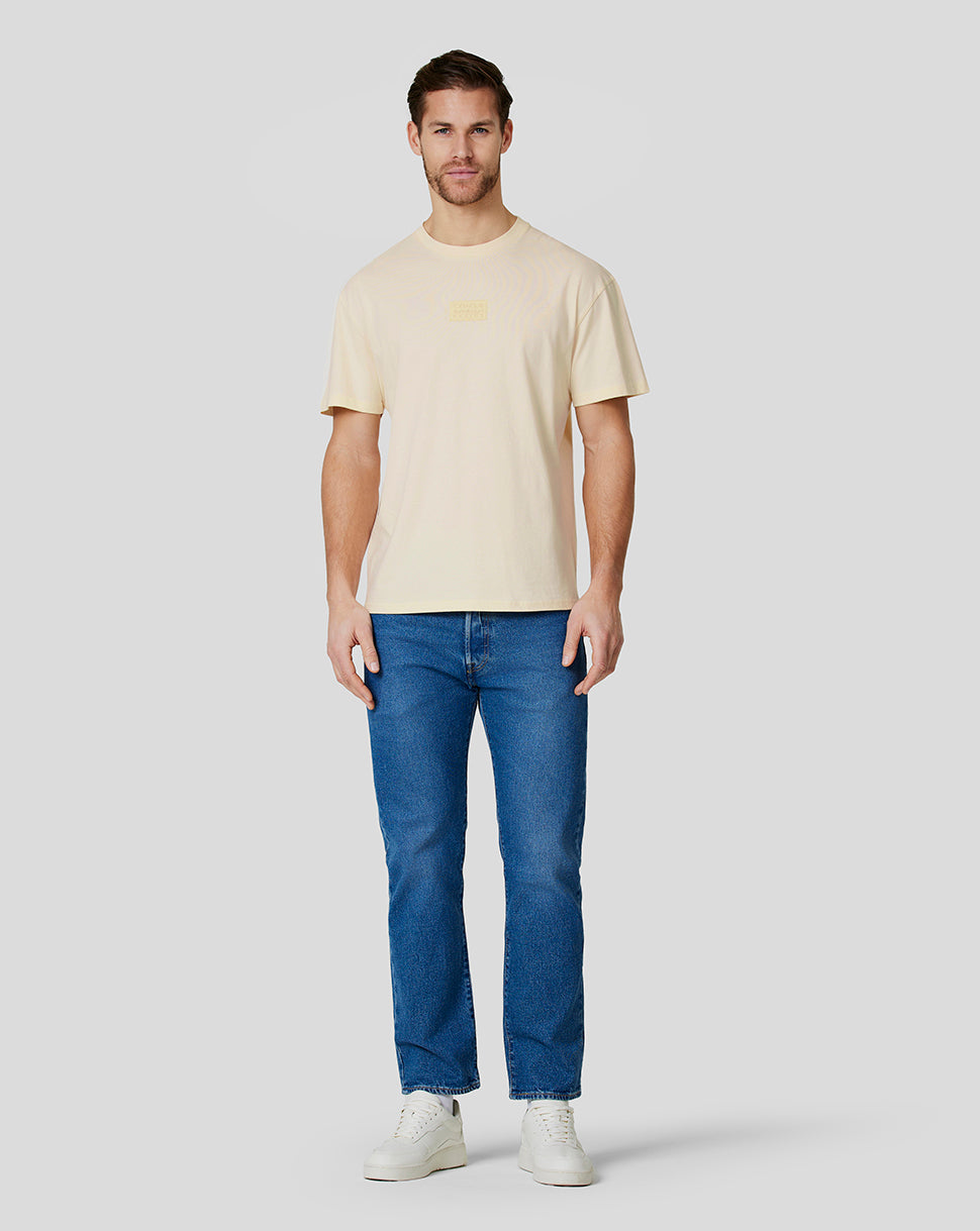 Full-length view of a man wearing a cream oversized T shirt with a chest patch logo, blue jeans and white sneakers
