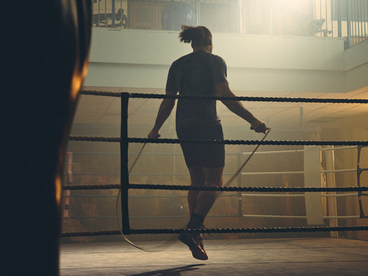 Boxer seen from behind inside a boxing ring, wearing a dark shirt with ropes visible in the foreground.