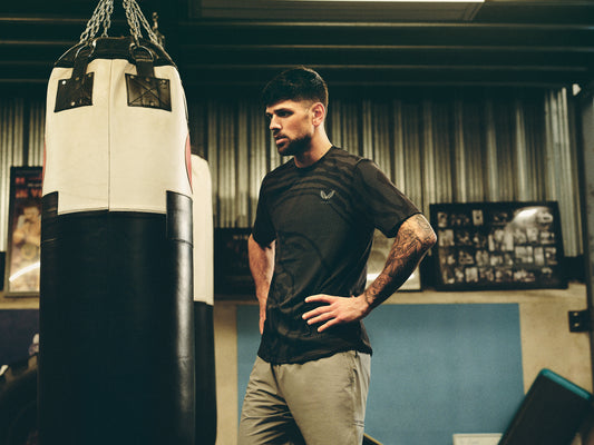 Athlete in a camo patterned shirt and beige shorts trains near a hanging punching bag in a gym