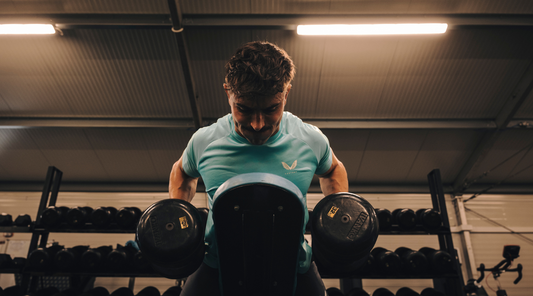 Athlete in a mint teal athletic tee performs a dumbbell bench exercise in a gym during pre-season.