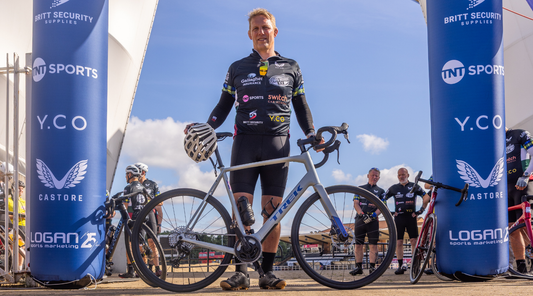 Male cyclist in black racing kit stands beside his gray Trek bike under blue sponsor arches at a race venue