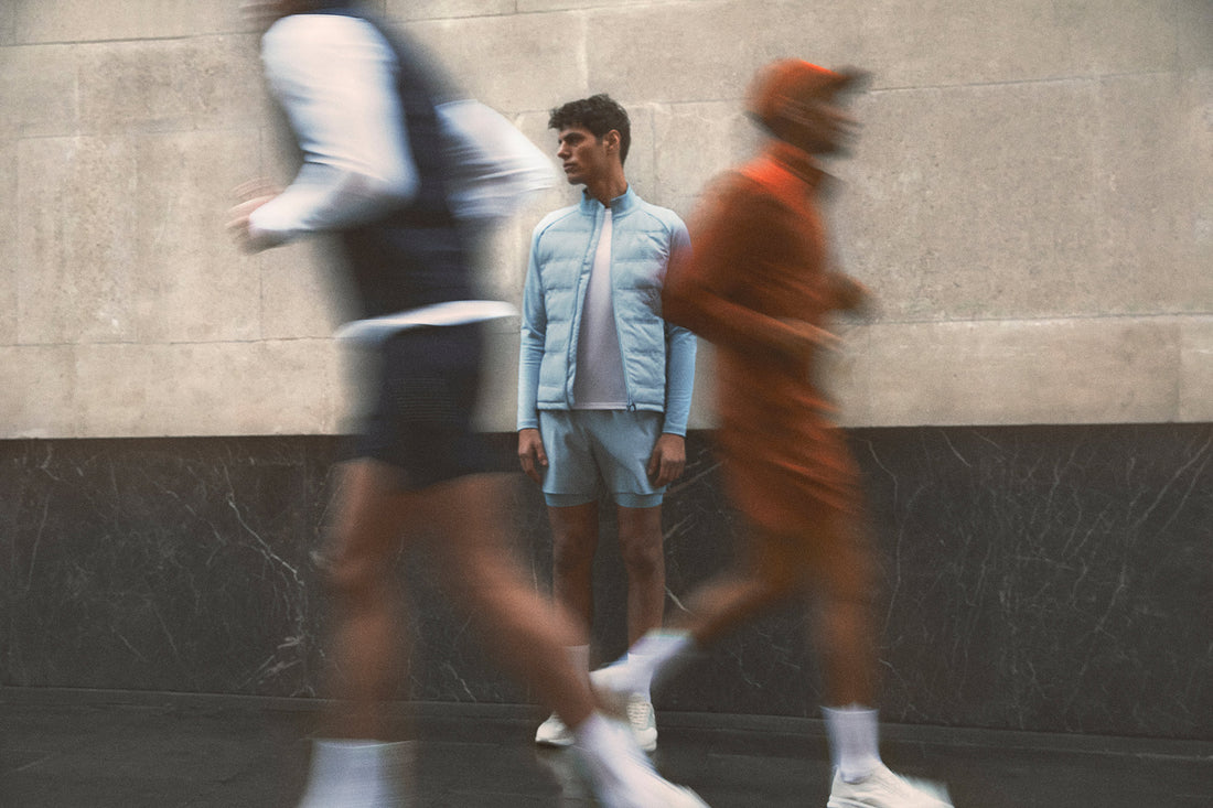 Model in pale blue padded jacket and shorts from Reiss x Castore SS24 stands against a light stone wall as blurred passersby move by.