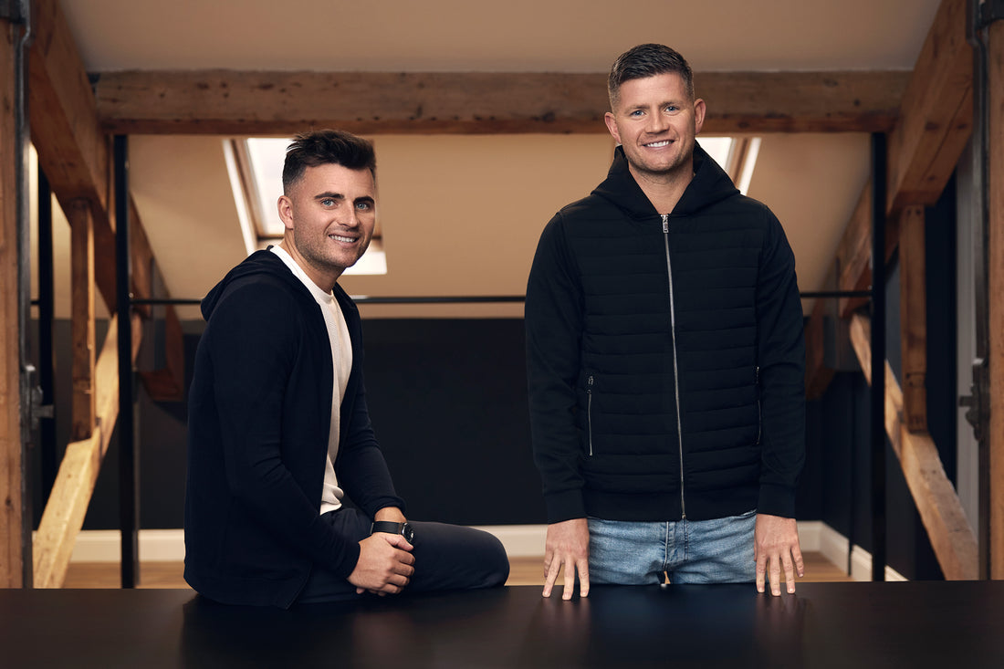 Two men in a wood framed studio lean on a table, one seated and the other standing, smiling.