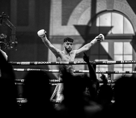 Black and white action shot of a muscular boxer in the ring with arms raised in victory behind branded ropes