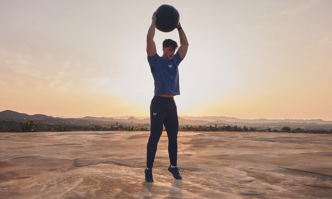 Athlete in blue training top lifts a large medicine ball overhead on a sunlit outdoor platform.