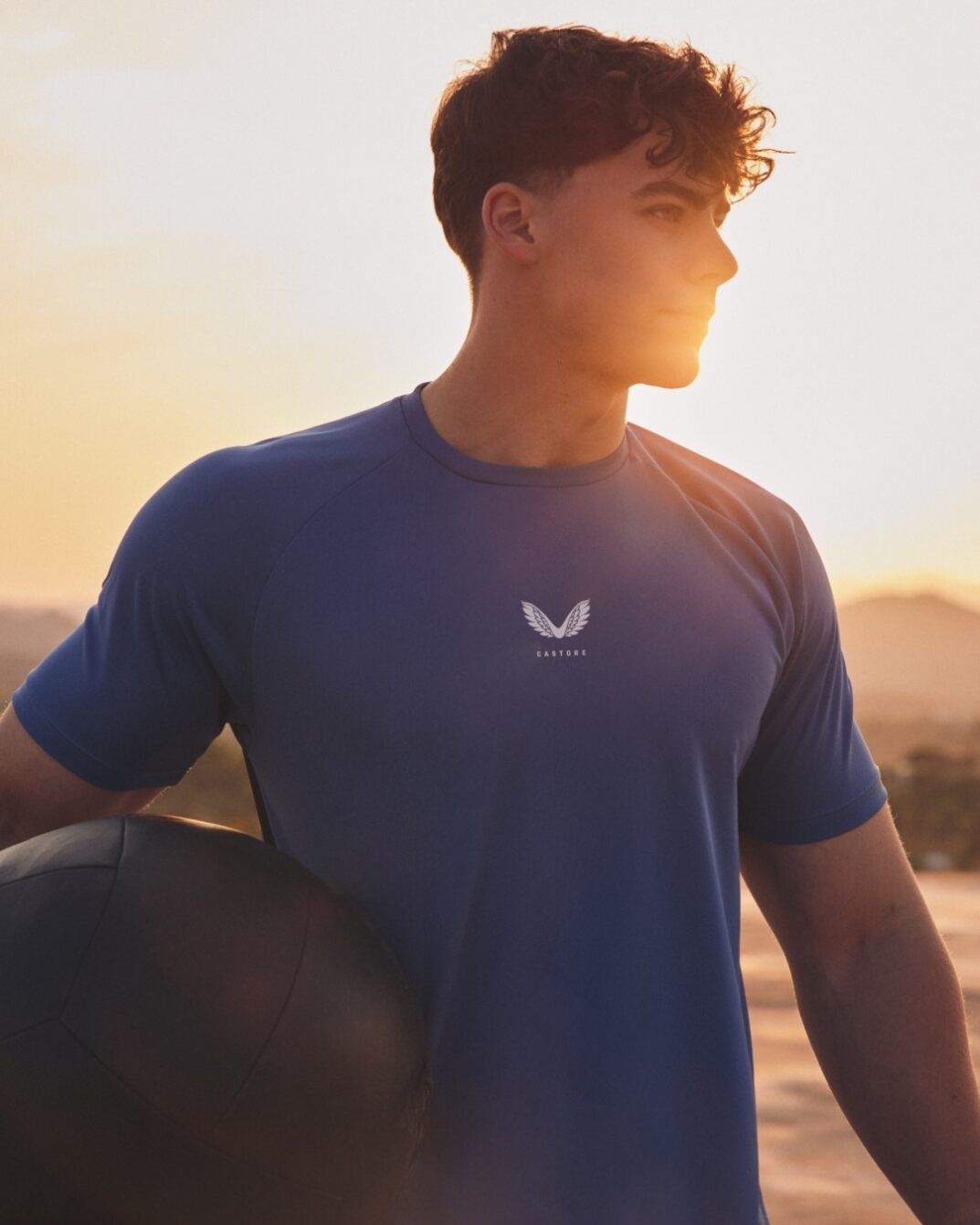 Athlete wearing a blue Castore shirt stands at sunset holding a large black ball