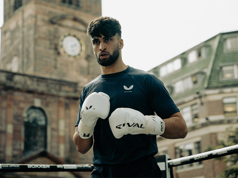 Young boxer in a navy shirt and white gloves stands in an outdoor boxing ring with a clock tower in the background.