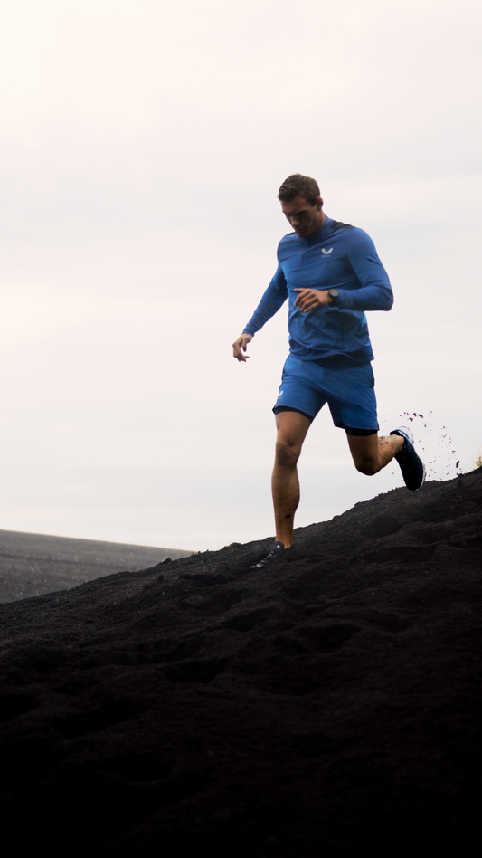 Man in blue running outfit sprinting up a dark volcanic hill under a pale cloudy sky.