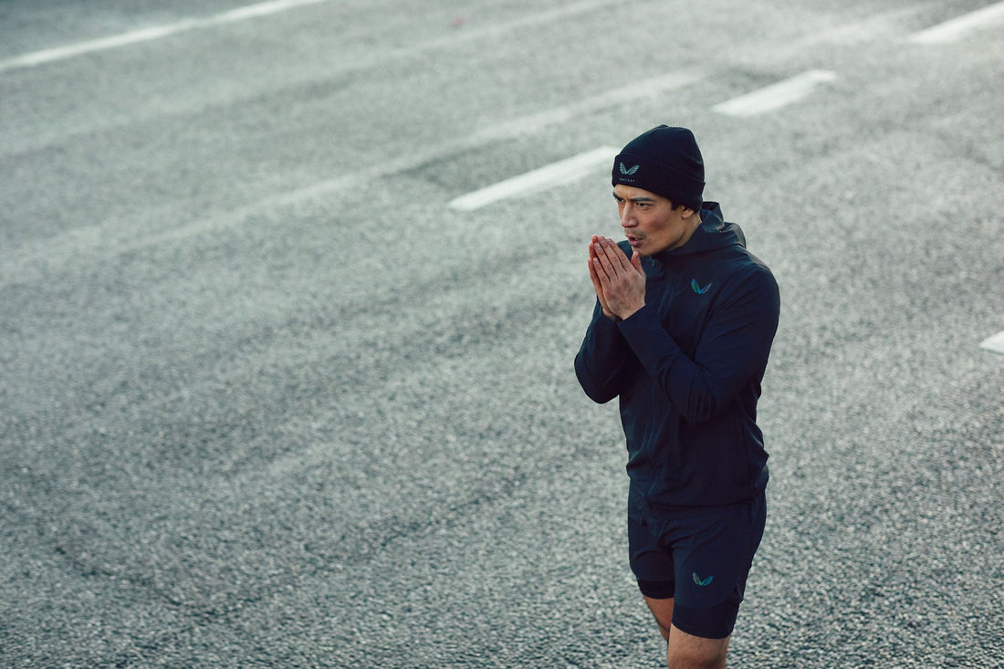 Athlete wearing a navy running jacket and beanie stands on a grey outdoor track with hands pressed together.