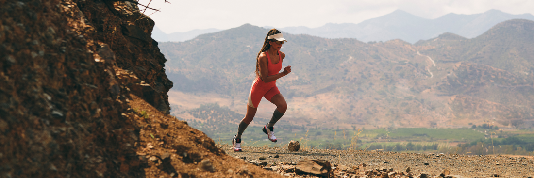 Athlete wearing coral Horizon running kit runs along a rocky hillside trail with distant mountains