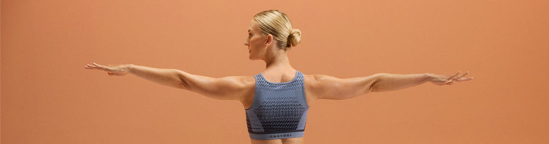 Woman in a blue patterned sports bra extends arms in a yoga pose against an orange backdrop