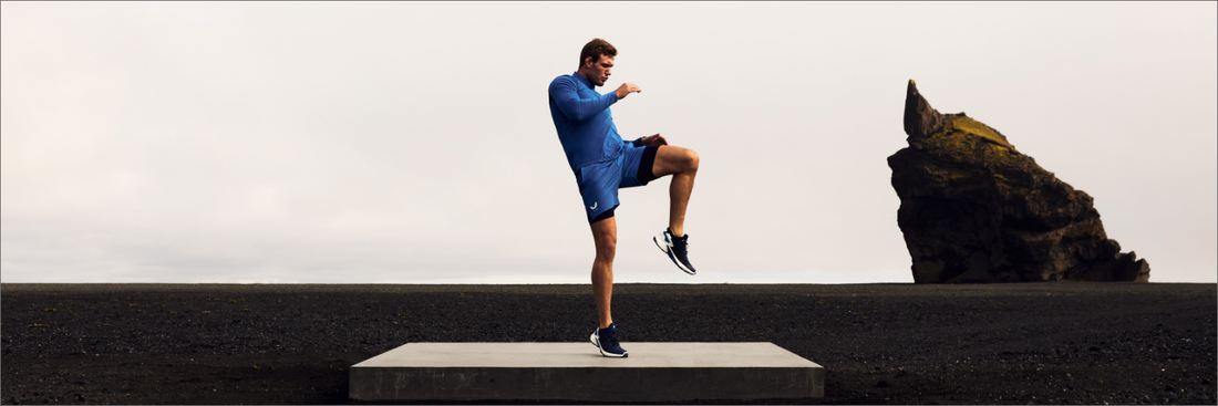 Male athlete in blue kit performs a high knee balance on a concrete platform in a stark coastal landscape