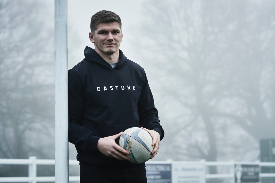 Man in Castore hoodie holding a rugby ball beside a vertical post outdoors.