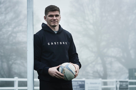 Man in Castore hoodie holding a rugby ball beside a vertical post outdoors.