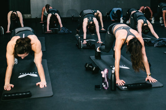 Group fitness class on mats doing planks in a dark gym with water bottles nearby