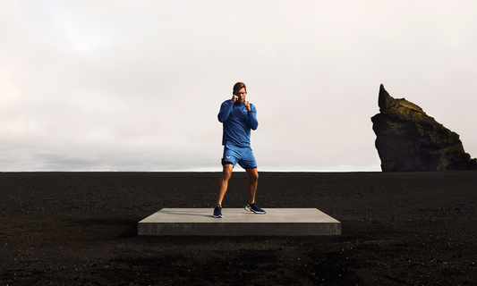 Runner in blue athletic wear on a concrete platform beside a black sand beach and a dramatic rock outcrop.