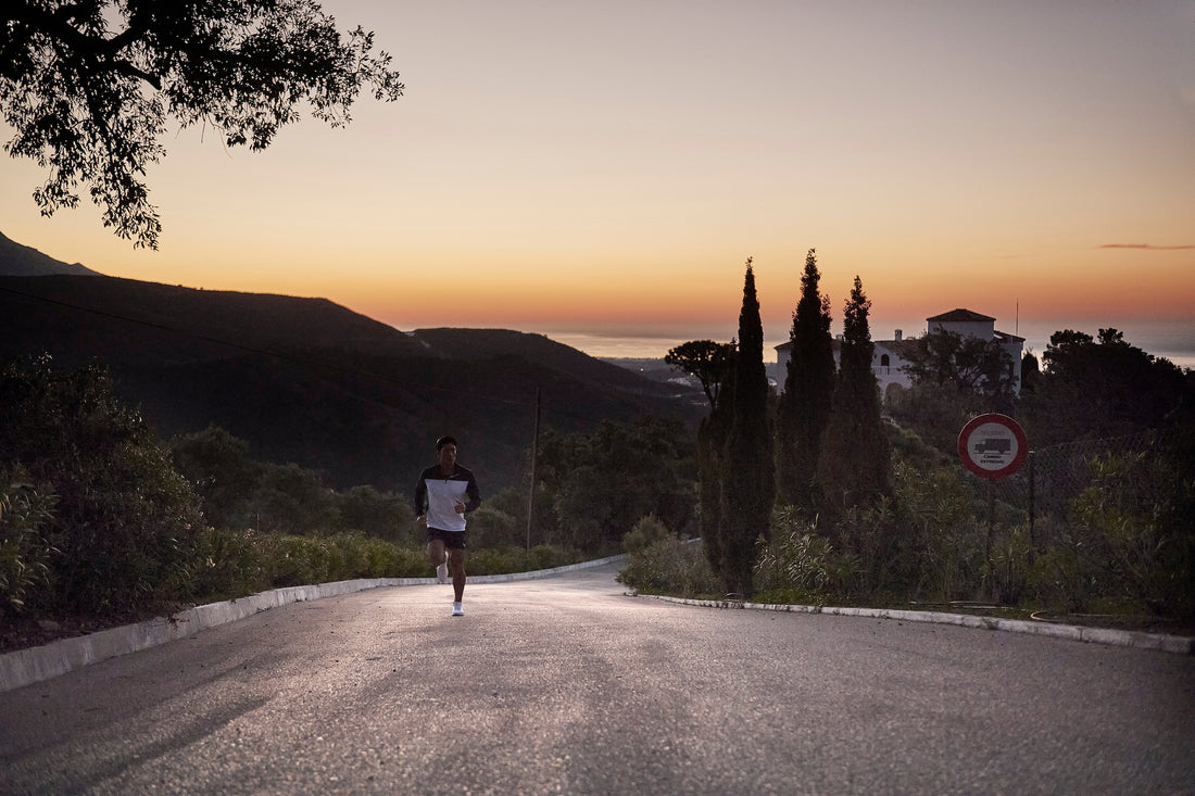 Runner in a white top and dark shorts on a road at sunset with silhouetted trees.