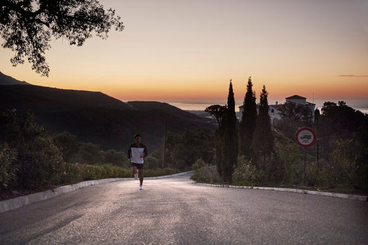 Runner in a white top and dark shorts on a road at sunset with silhouetted trees.