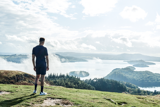 Man stands with his back to the camera on a hilltop overlooking a misty lake and rolling hills.