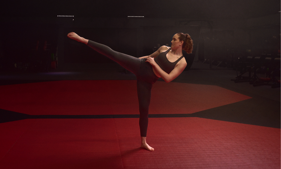 Athlete in a black sports top and leggings demonstrates a high kick on a red gym mat during a studio workout.
