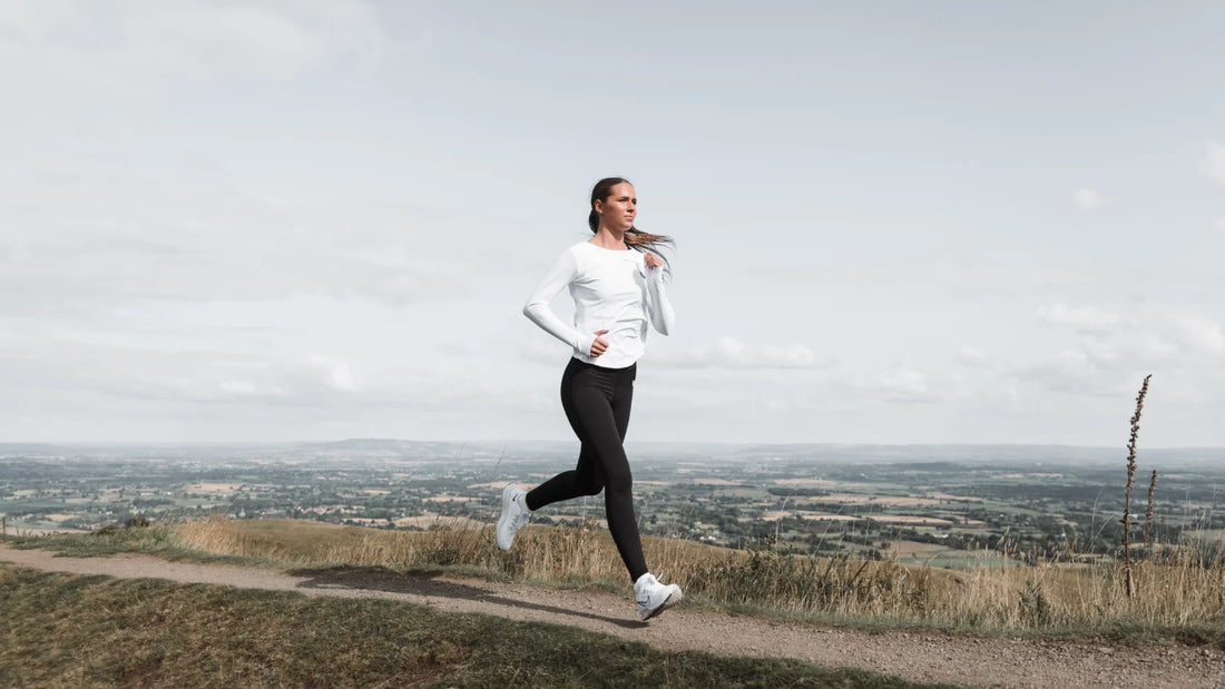 Woman in white long sleeve top and black leggings runs along a hillside trail with a wide rural valley in the background.