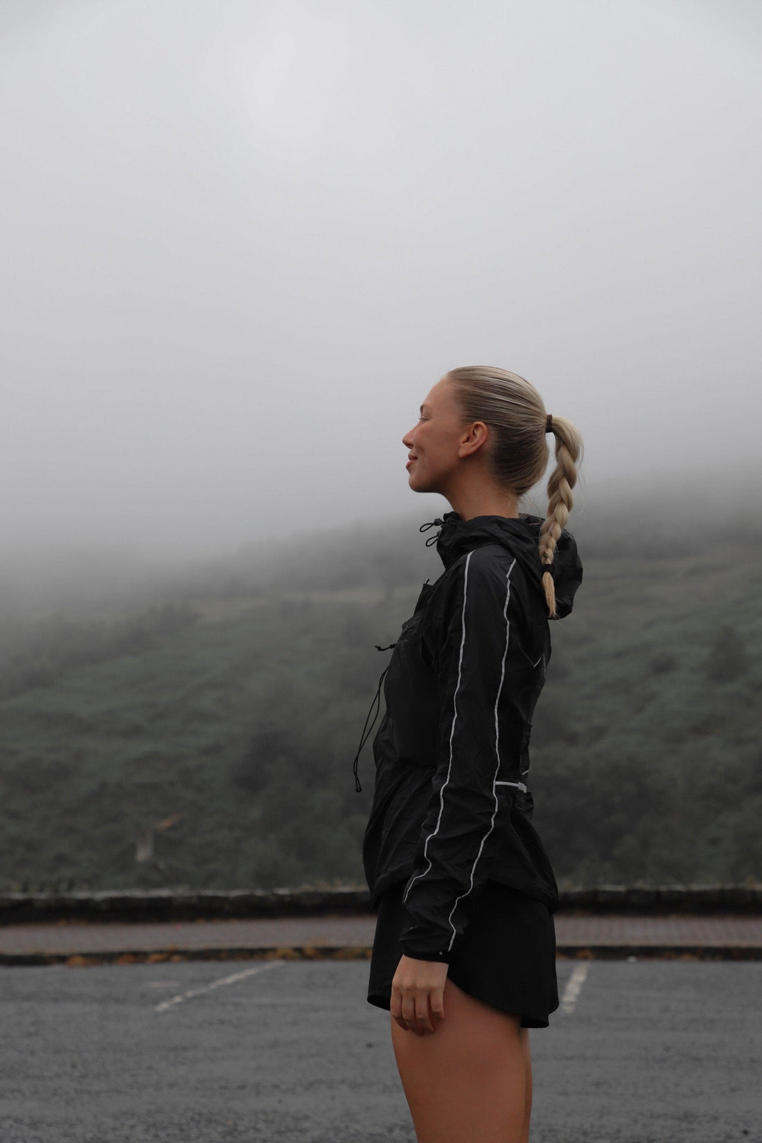 Side profile of a female runner with a long blonde braid wearing a black waterproof jacket on a misty hillside