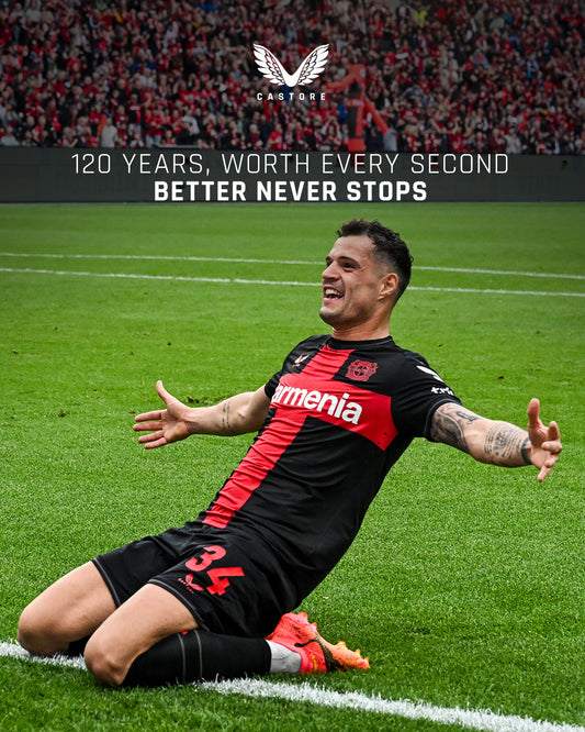 Bayer Leverkusen footballer celebrates on the pitch in a black and red kit as fans cheer in the stands.