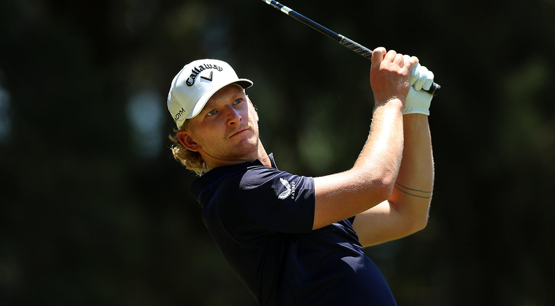 Golfer in a white Callaway cap and navy polo mid backswing with a club against a blurred green background.