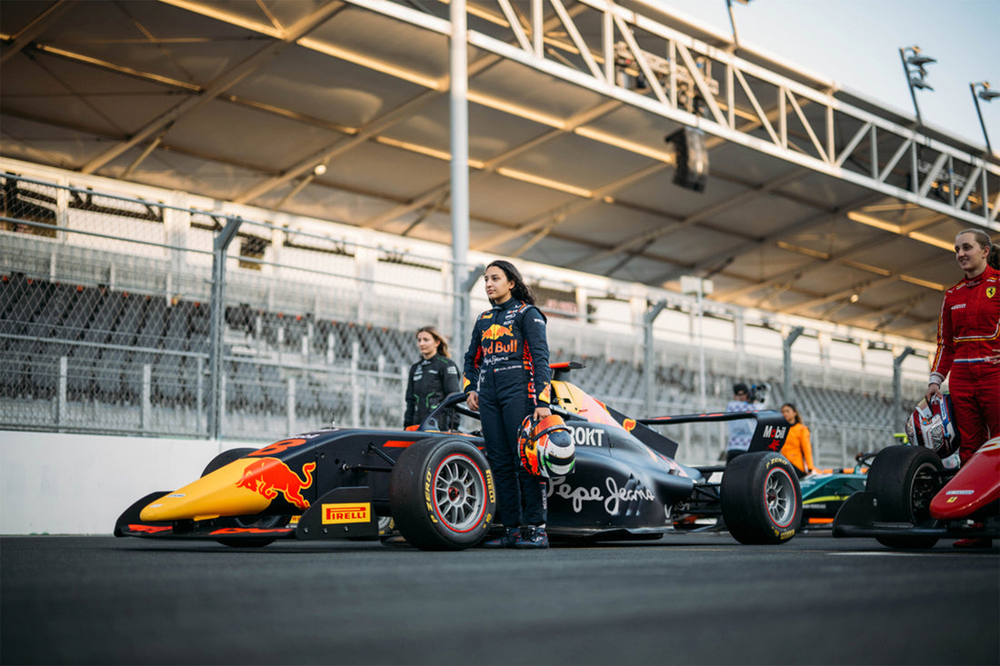 Female driver in Red Bull racing suit stands beside a Formula 1 car in the pit lane.