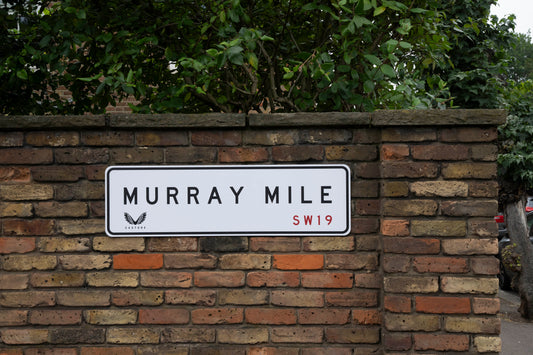 Murray Mile street sign mounted on a brick wall with green shrubbery and trees in the background.
