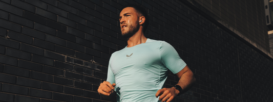 Male runner in a light blue athletic shirt sprints past a dark brick wall while wearing wireless earbuds.