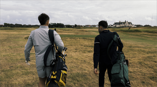Two golfers walk across a windswept course carrying large golf bags one in a light gray jacket the other in dark gear with houses in the distance