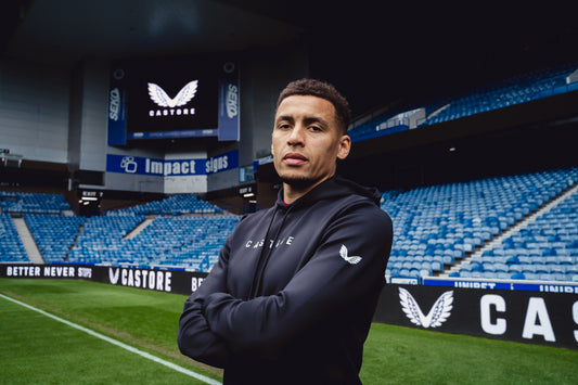 Portrait of a man in a dark hoodie at a stadium with blue seats and a large screen displaying the Castore logo.