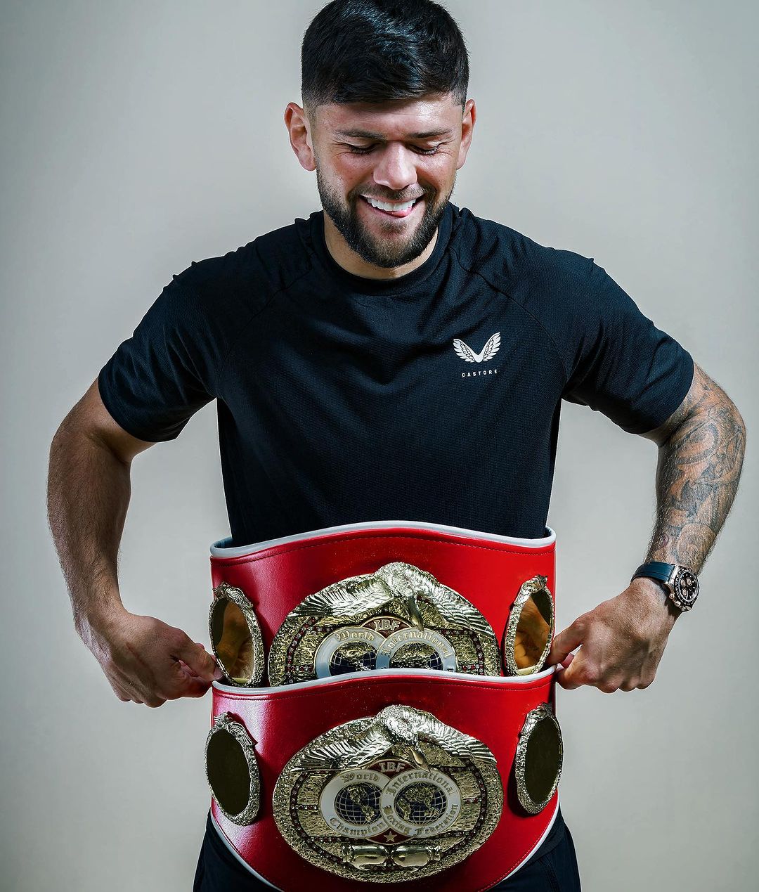 Smiling boxer with a large red world championship belt featuring gold plates around his waist