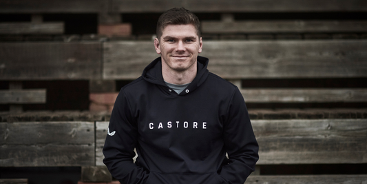 Athlete wearing a black Castore hoodie sits on weathered wooden steps outdoors.