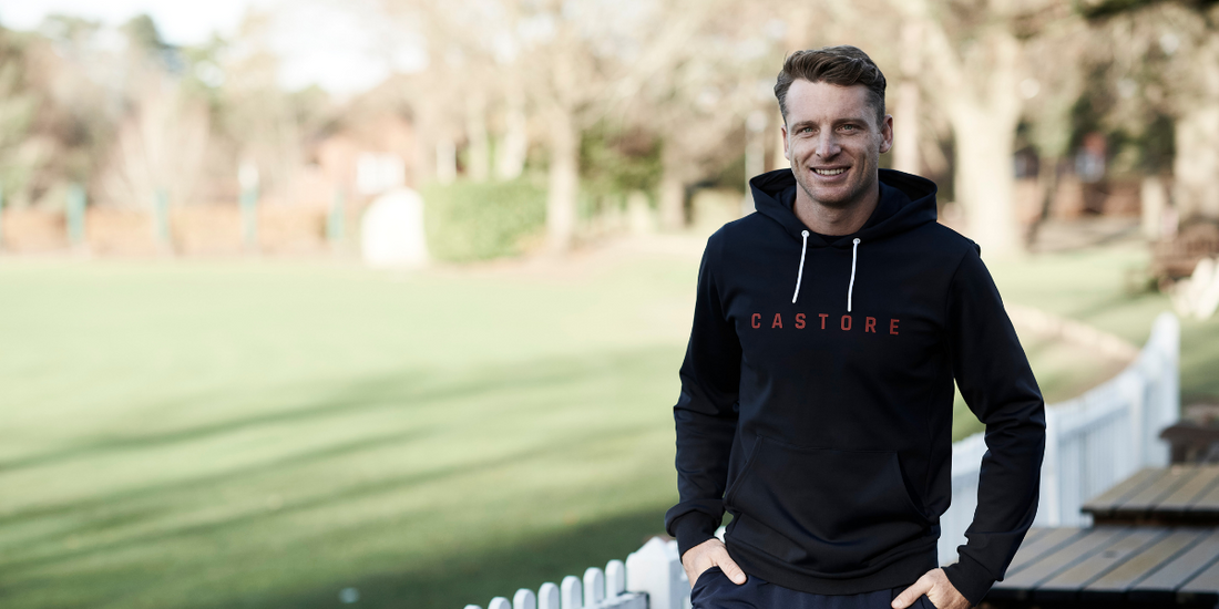 Smiling man in a black Castore hoodie with red logo standing outdoors near a fence.