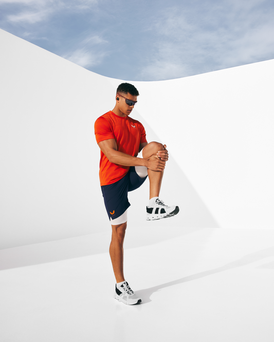 Male model in bright orange Castore kinetic collection top with navy shorts against a white backdrop